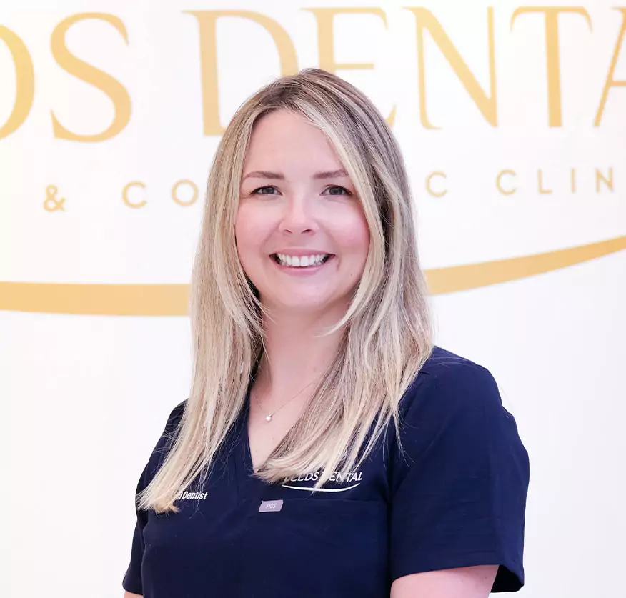 A smiling female dentist in a Leeds Dental uniform stands confidently in front of the clinic’s logo.