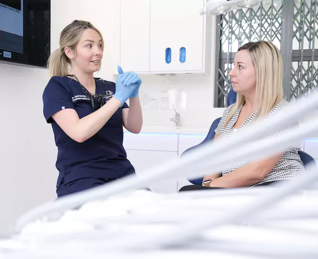A dentist wearing blue gloves explains a treatment plan to a patient during a TMJ consultation.