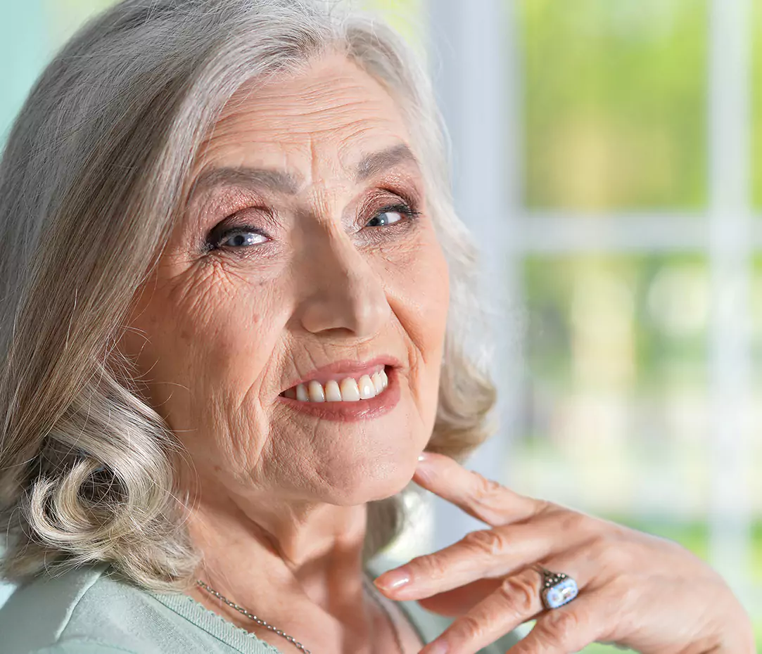 Elderly woman with silver hair smiling confidently, showing healthy teeth, while posing in a bright indoor setting.