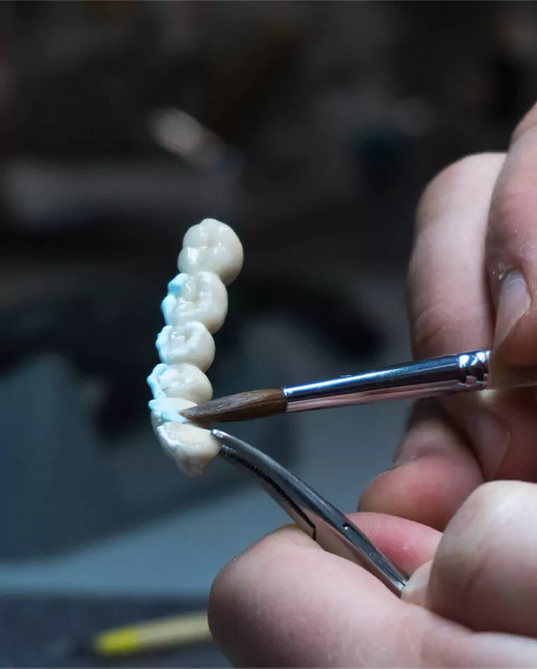 Dental technician carefully shaping a dental bridge with a fine brush and tweezers.