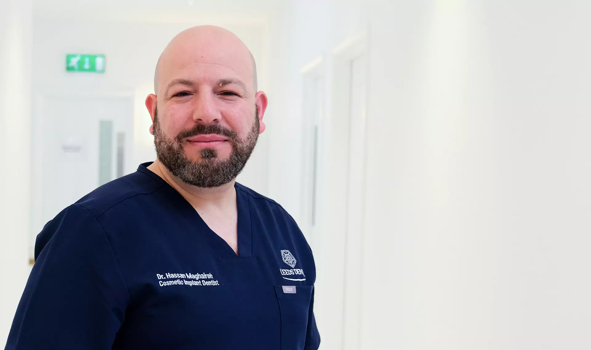 Dr. Hassan Maghaireh, a cosmetic implant dentist at Leeds Dental, wearing a navy blue uniform, standing in a bright clinic hallway.