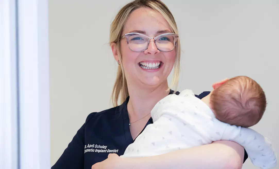 Dr. April Scholey, a cosmetic implant dentist, smiling while holding a baby.