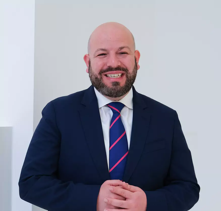 A smiling bald man with a beard wearing a navy-blue suit and striped tie stands confidently against a plain background.