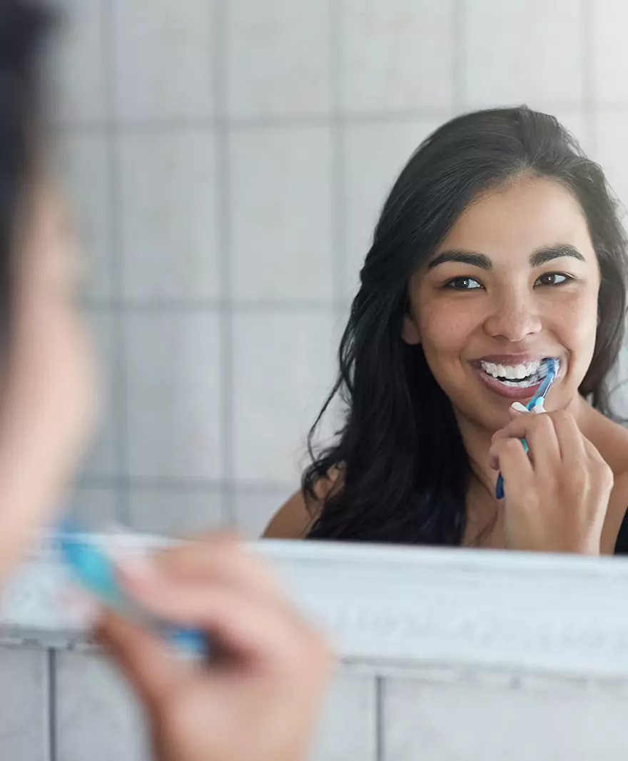 A smiling woman brushing her teeth in front of a mirror.