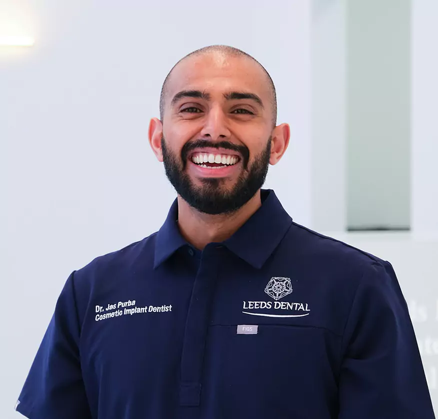 A smiling male dentist in a Leeds Dental uniform stands confidently in a modern clinic setting.