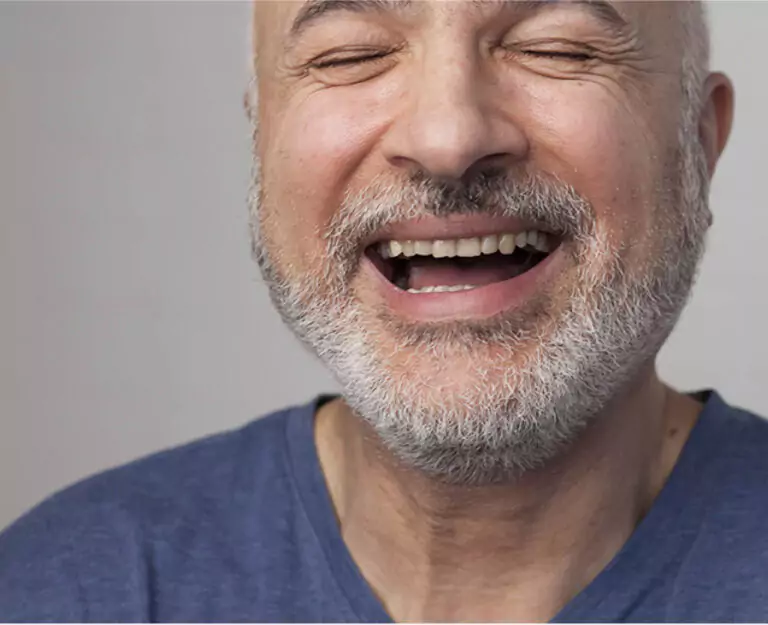 A bearded man in a blue shirt laughing with his eyes closed, showing a bright and confident smile.
