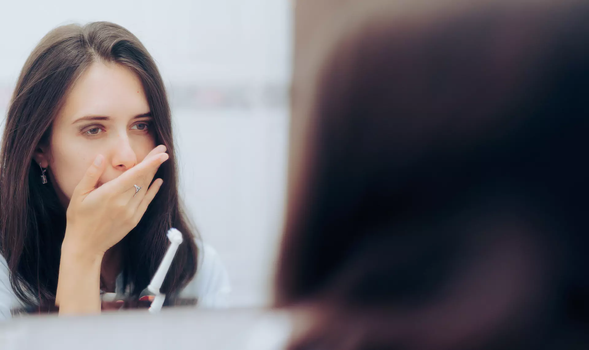 A woman covering her mouth while looking in the mirror, appearing worried about her breath or dental health.