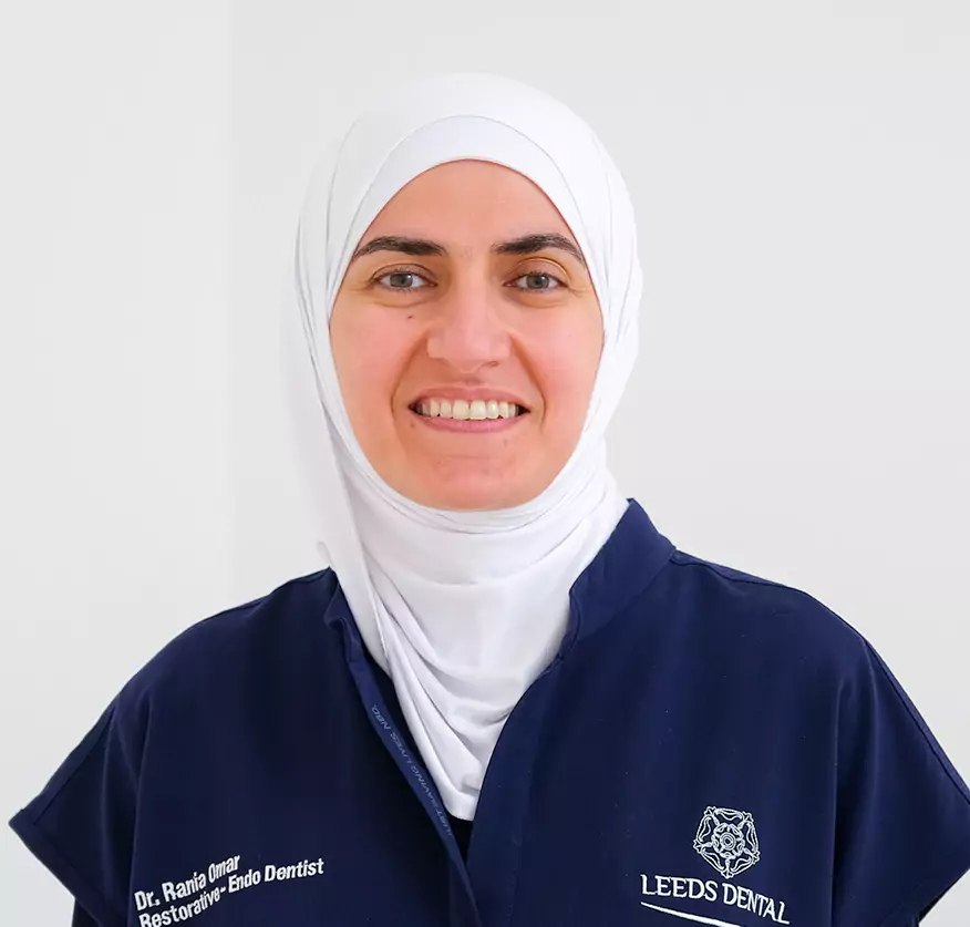 A smiling female dentist wearing a hijab and a Leeds Dental uniform stands confidently against a plain background.