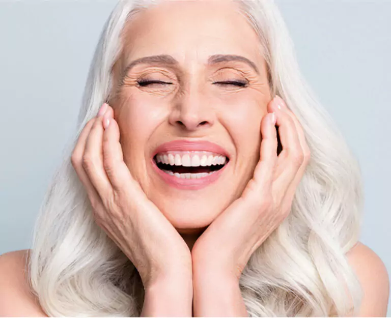 A joyful older woman with silver hair smiling brightly and holding her face in happiness.
