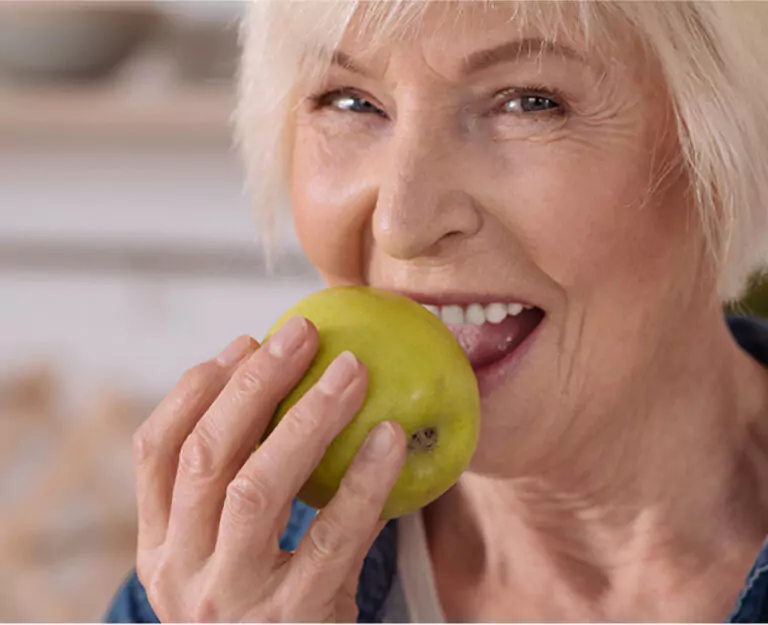 An elderly woman with white hair smiling as she takes a bite of a green apple, showcasing her strong teeth.