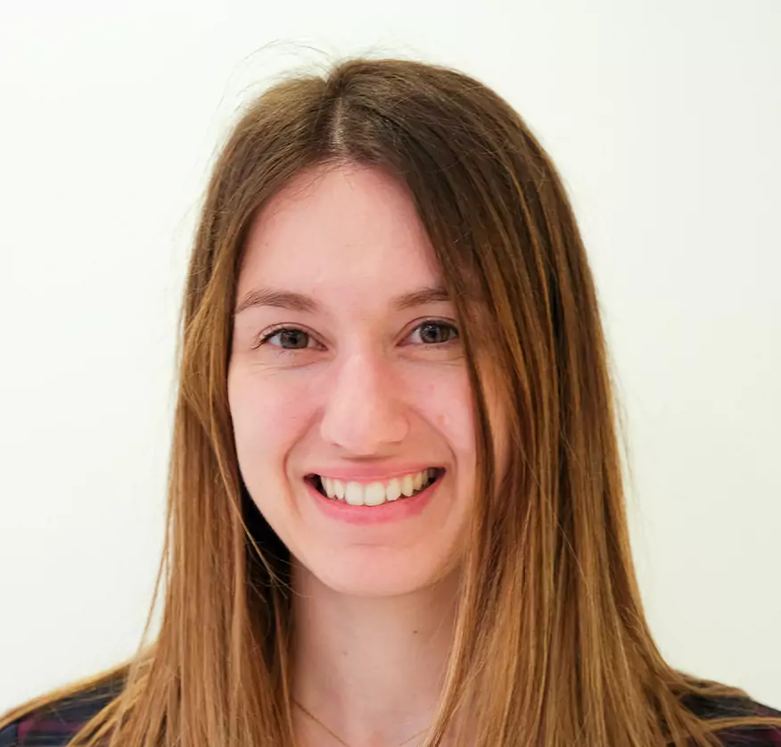 A smiling female dentist with long brown hair poses confidently against a plain background.