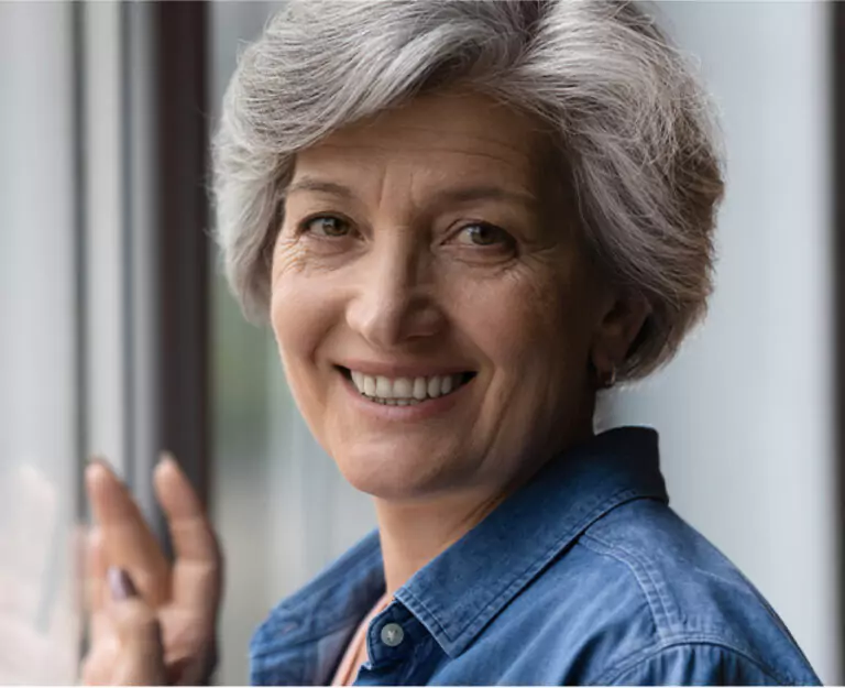 A mature woman with short silver hair, wearing a denim shirt, smiling warmly by a window.